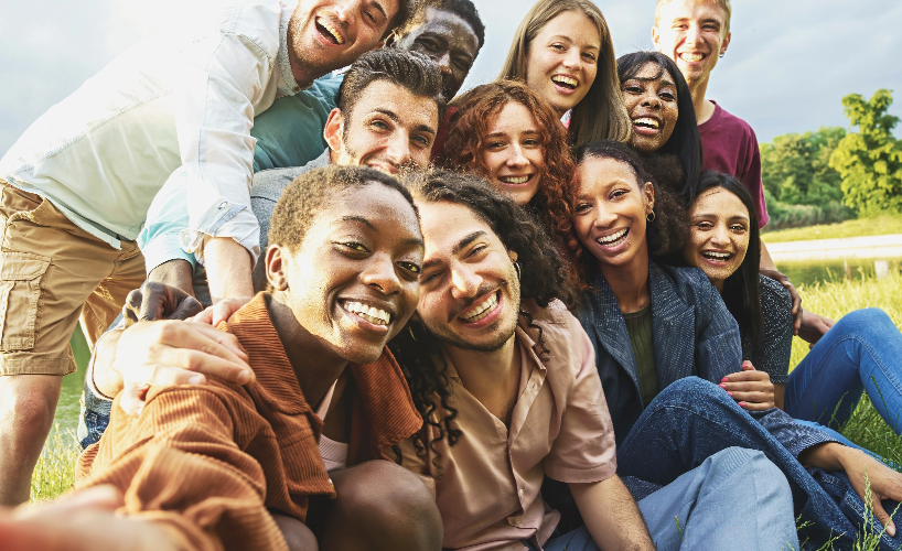 A group of diverse and smiling young adults taking a selfie outdoors in a sunny, grassy area.